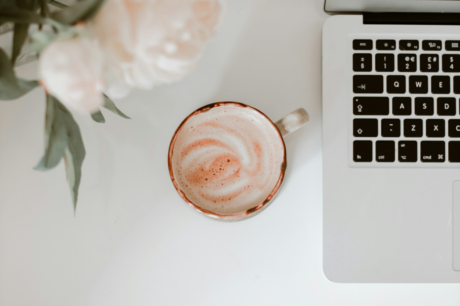 Um café com leite ao lado do teclado de um portátil e peónias rosa-claro