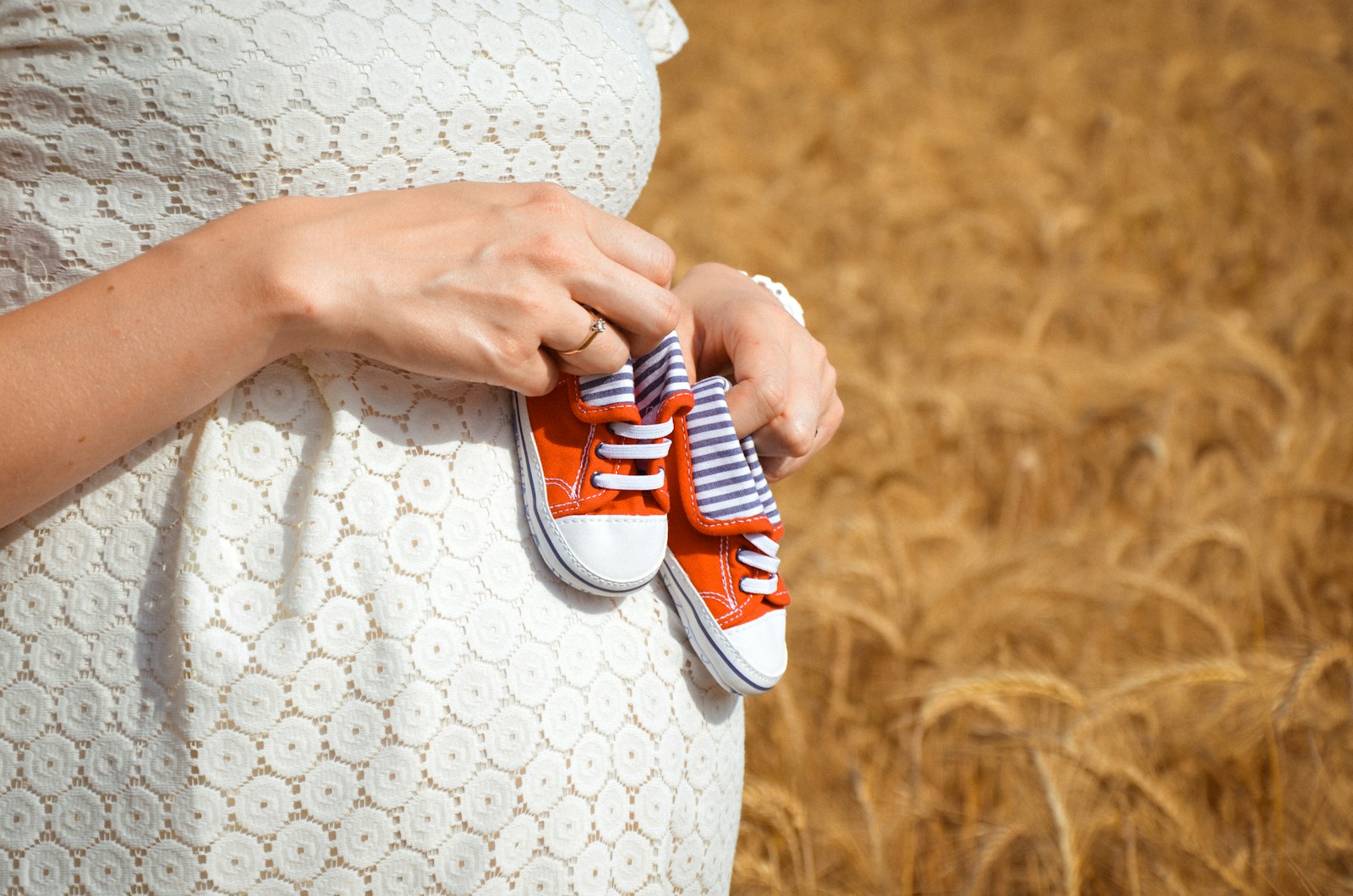 Schwangere Frau im weißen Spitzenkleid hält winzige Babyschuhe vor einem Weizenfeld