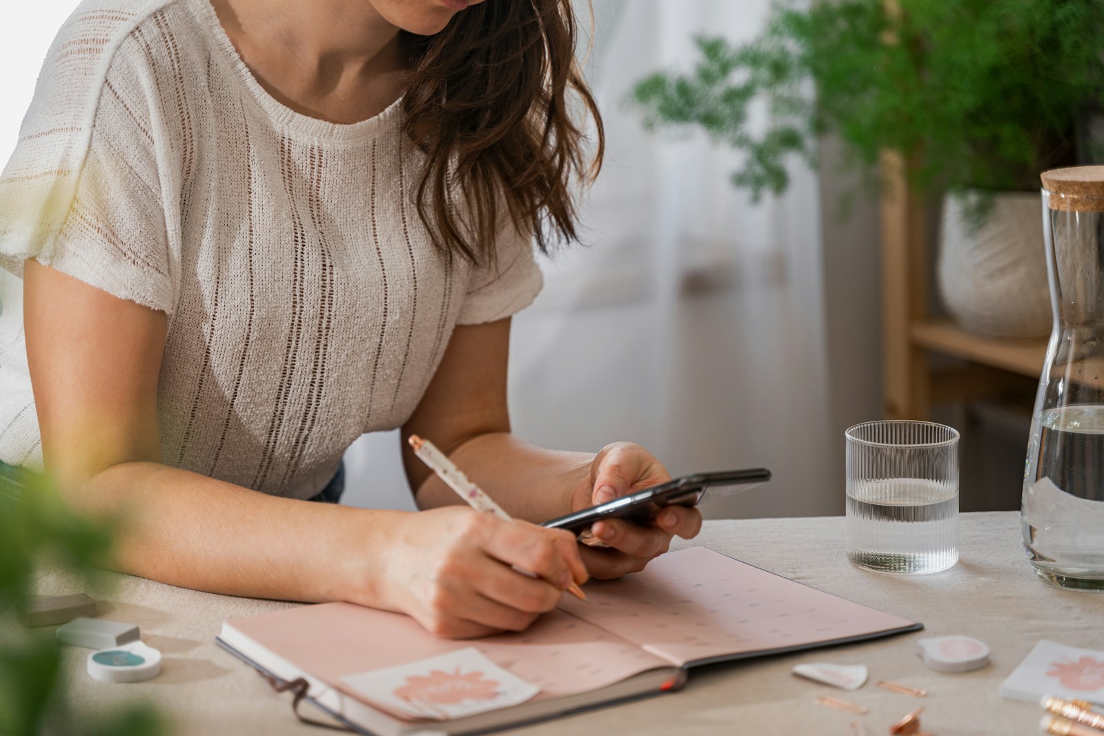 Uma mulher escreve numa agenda cor-de-rosa à mesa, com o telemóvel na mão