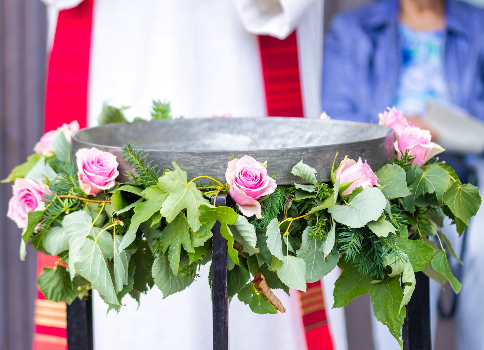 Taufbecken mit rosa Rosen und Grün geschmückt, dahinter der Talar des Pfarrers