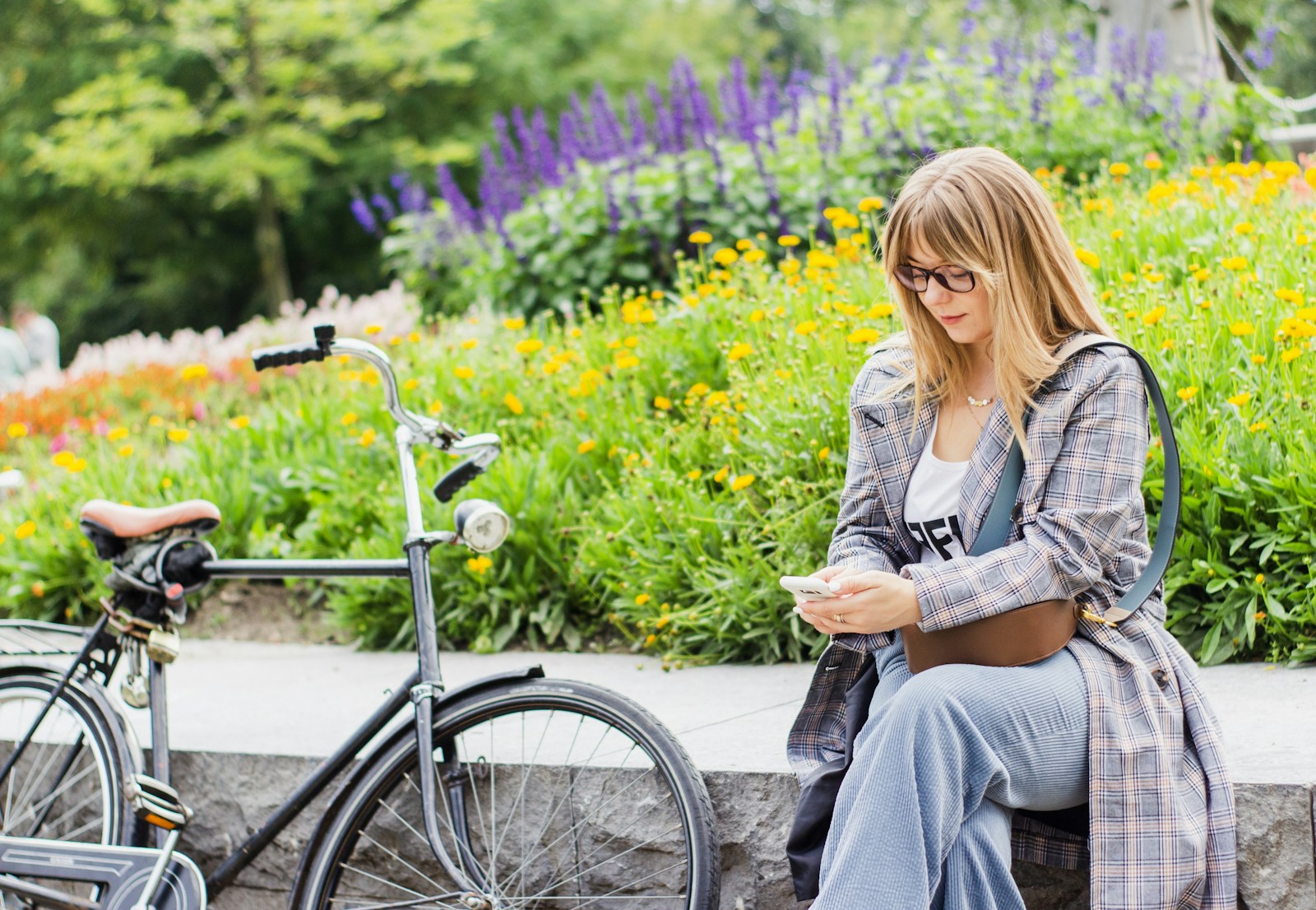 Frau mit Brille liest auf ihrem Handy, neben Blumenbeeten und einem Fahrrad