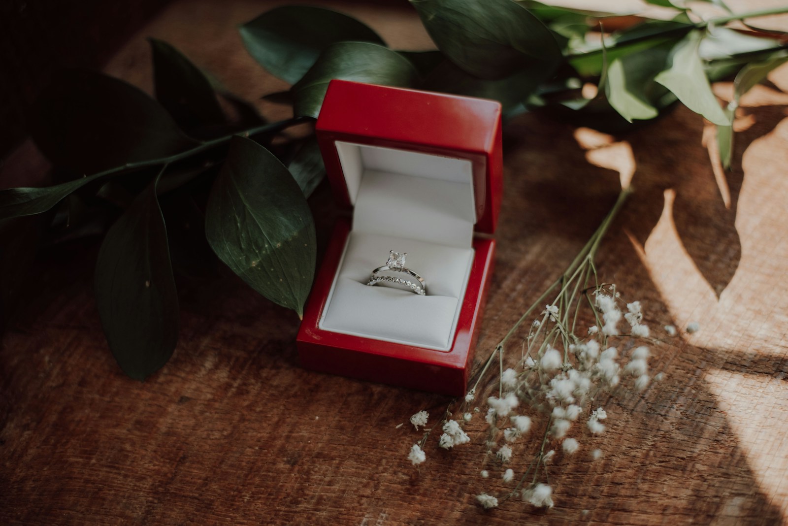Bague de fiançailles et alliance dans un écrin rouge ouvert sur du bois ensoleillé