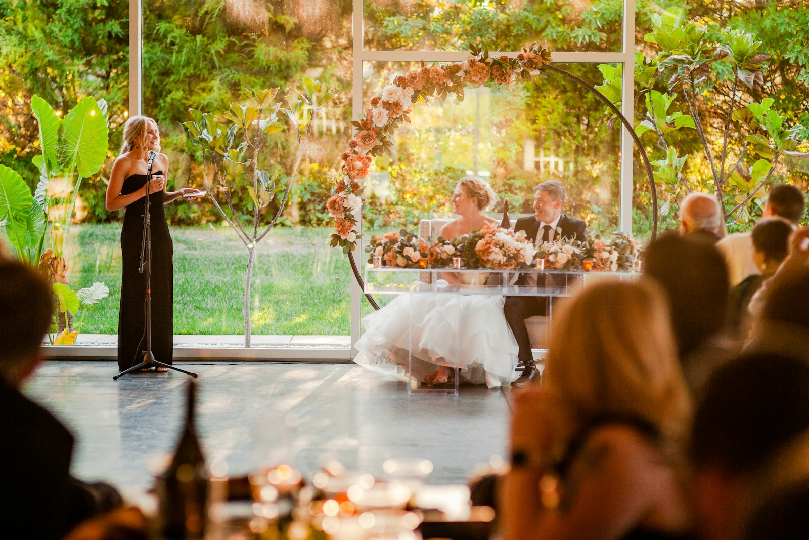 A woman speaks at a microphone while the newlyweds listen from their flower-covered table