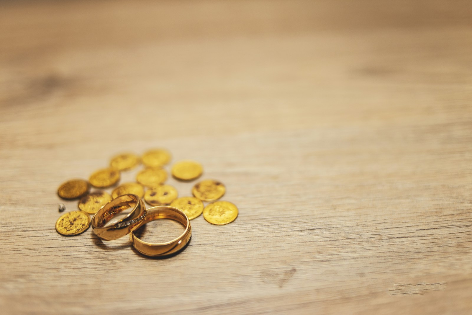 Twee gouden trouwringen naast een handvol gouden munten op licht hout
